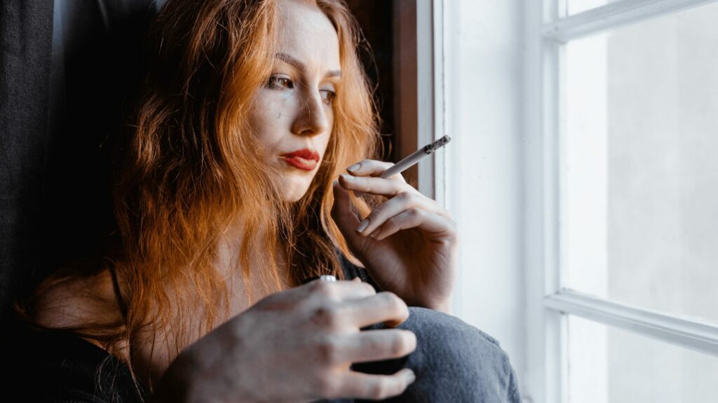 Femme songeuse fumant une cigarette devant une fenêtre, semblant réfléchir à l’arrêt du tabac et aux idées reçues, illustration des questionnements avant un accompagnement dans un centre LaserAddict Maroc.